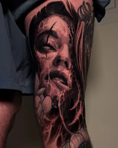 Dark surrealism thigh tattoo by Andrew Stopay: a female face with a scarred eye, organic flesh textures and flowers